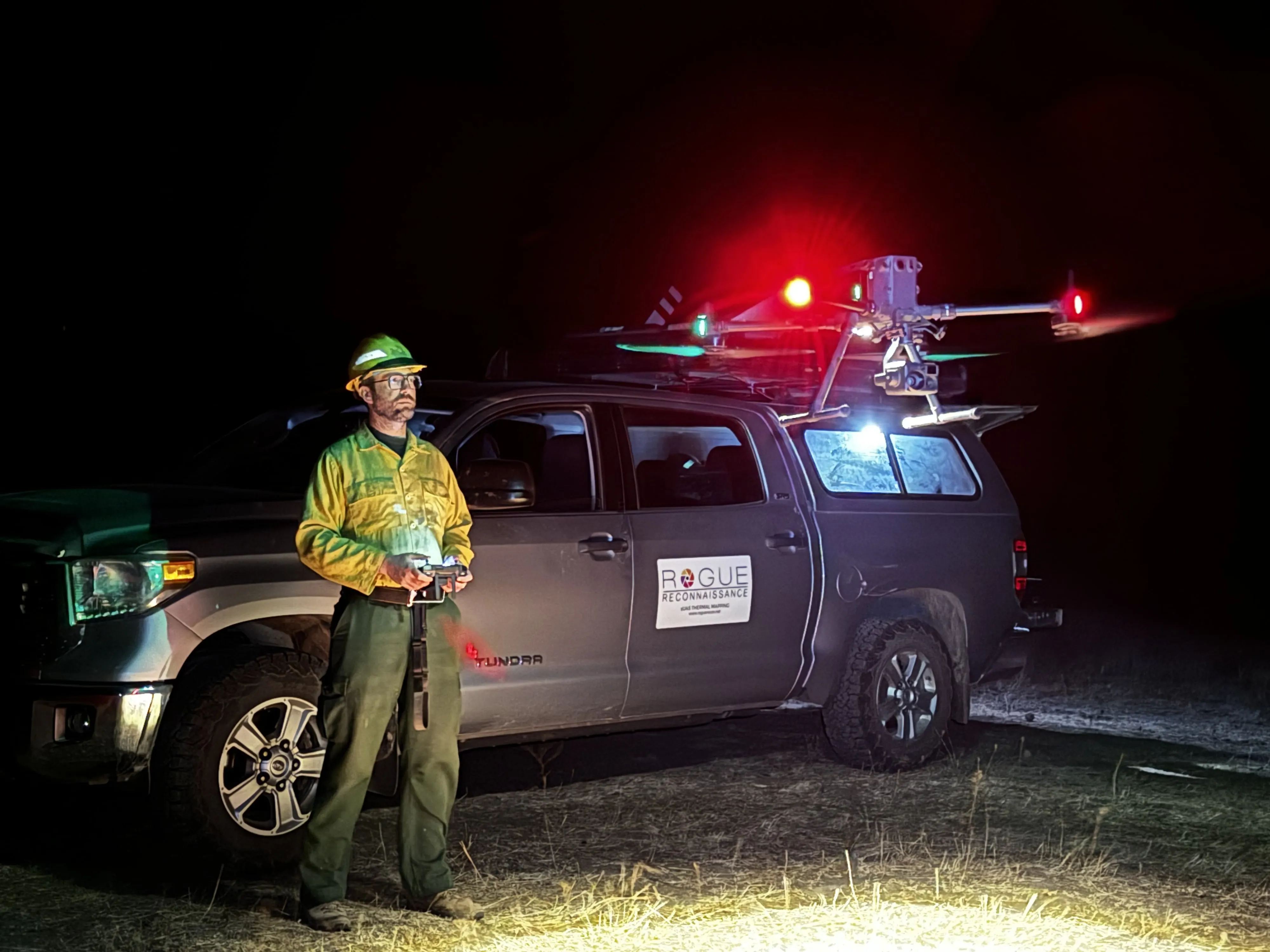 The aircraft lit up at night beside the Rogue Reconnaissance truck, the pilot holding the controller.