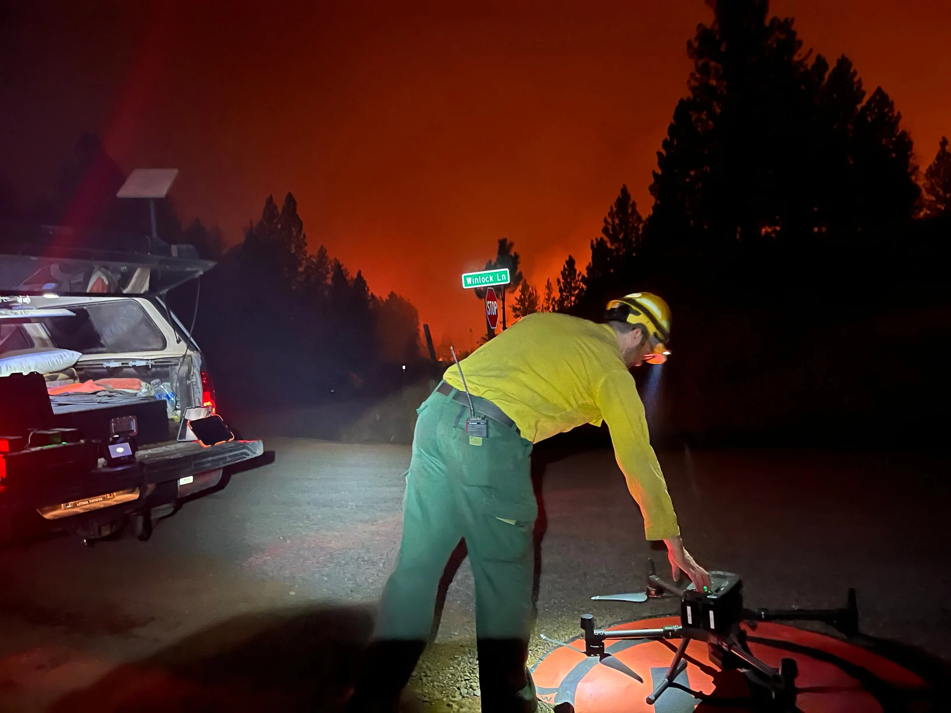 A pilot in a yellow fire shirt setting the aircraft on the truck tailgate at dusk, the sky red with smoke.