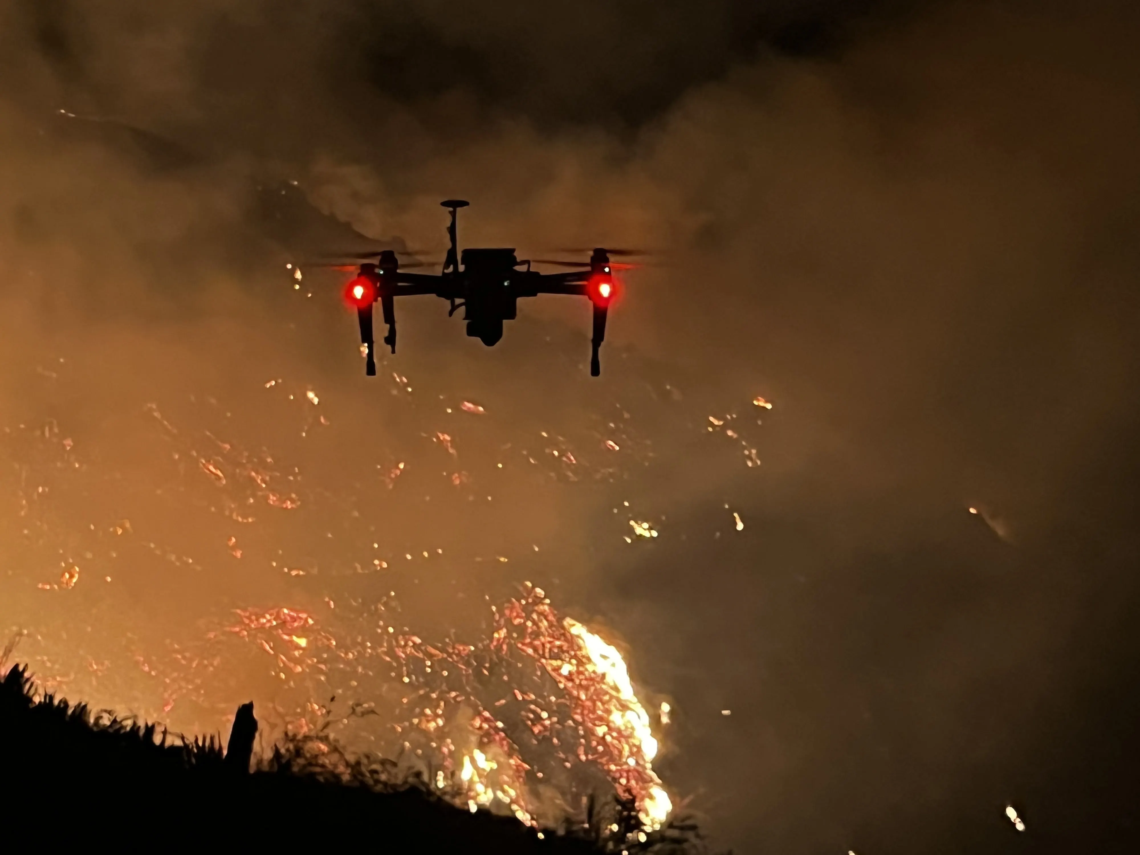 The aircraft holding over a burning hillside at night, embers on the slope and smoke rising past it.